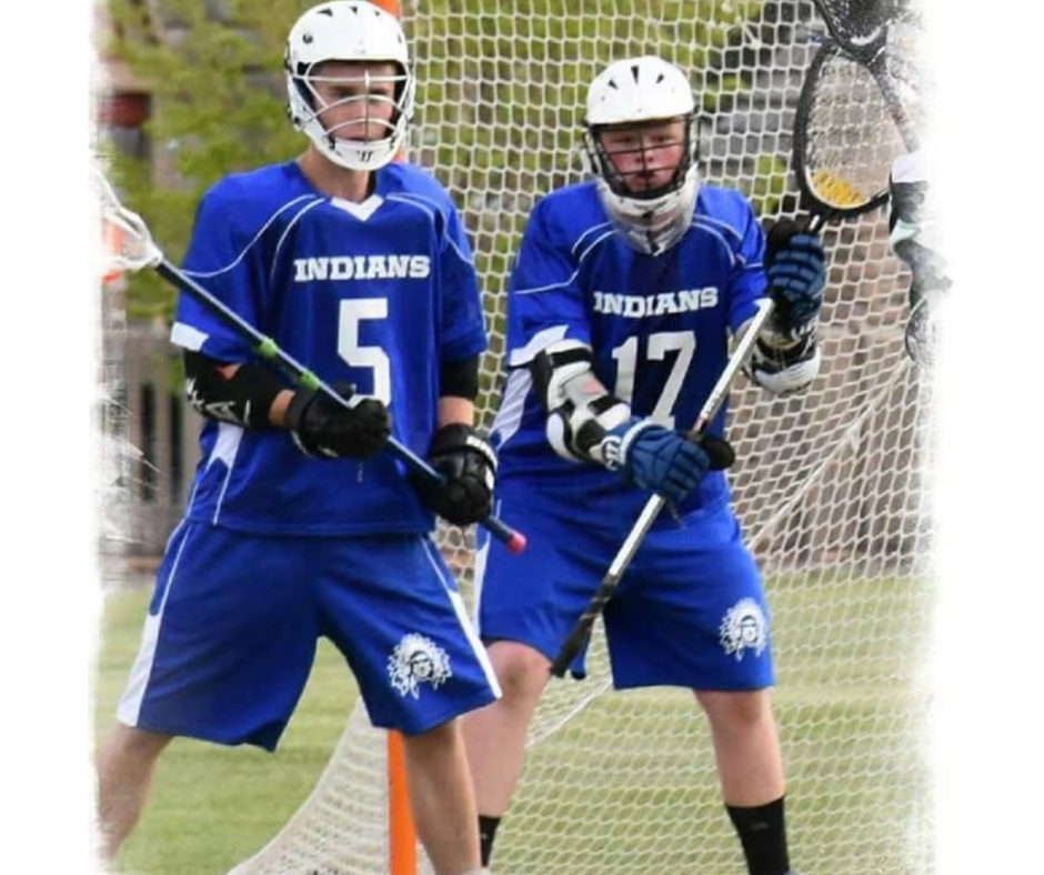Bailand, in lacrosse goalie gear, with a teammate in front of the goal.